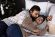 © Елена Бабанова - A man is sleeping with his arm around a woman in a bed. The woman is resting her head on a pillow, and the man is kissing her forehead. There is a Christmas tree in the background.