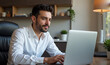 © dapra01 - A Hispanic man in his late twenties, wearing a crisp white button-up shirt and dark slacks, is seated at a stylish home office desk focused on his laptop