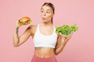 Young shocked woman she wear white top casual clothes hold eat choose between lettuce salad and burger isolated on plain pink background. Proper nutrition healthy fast food unhealthy choice concept.