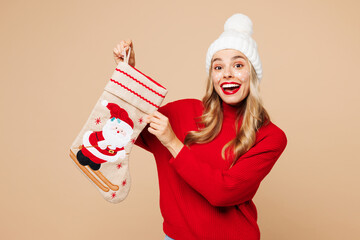 Canvas Print - Merry young surprised fun woman wear red warm cozy sweater hat posing hold in hand stocking sock isolated on plain pastel light beige background. Happy New Year celebration Christmas holiday concept.