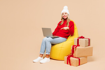 Wall Mural - Full body merry young IT woman she wears red sweater hat posing sit in bag chair hold use work point on laptop pc computer isolated on plain beige background. Happy New Year Christmas holiday concept.