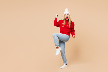 Full body merry young woman wear red warm Christmas sweater hat posing do winner gesture celebrate clench fists say yes isolated on plain beige background. Happy New Year celebration holiday concept.