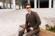 © dikushin - Portrait of stylish photogenic young businessman in brown suit and sunglasses sitting confidently on city bench, exuding professionalism and success in modern urban setting. Concept of business life.