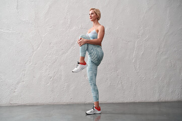 Female athlete warming up in the gym. Caucasian sportswoman standing on one leg, the other leg bent at the knee and pressed to the chest.