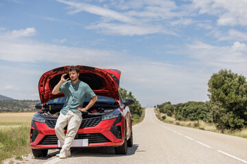 Young overwhelmed and angry man on vacation with reflective vest and broken car on the shoulder calling insurance on the phone, requesting a tow truck.