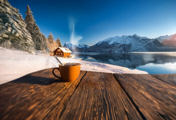 Poster - A steaming cup of coffee sits on a wooden table overlooking a snowy lake and mountains with a small wooden cabin in the background.
