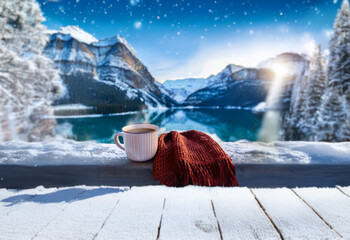 Poster - A mug of hot beverage rests on a wooden railing, covered in snow, with a cozy scarf and a breathtaking view of a snow-capped mountain range and a serene lake.