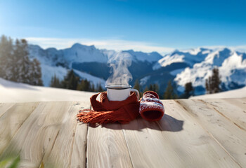 Poster - A steaming cup of coffee on a wooden table with a red scarf and mittens in front of a snowy mountain landscape.