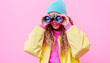 © Poj - A beautiful woman wearing colorful dress holding binocular on two hands with against pink background.