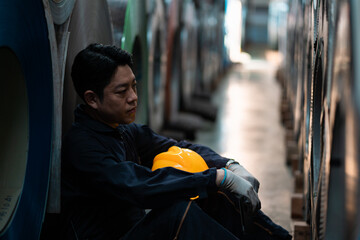 A man in a yellow helmet sits on the floor in a factory. He is wearing a black shirt and blue gloves