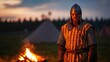 © GreenMOM - A warrior stands by a campfire, showcasing medieval attire against a twilight backdrop of tents and trees.