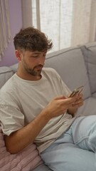 Wall Mural - Young man smiling while using smartphone in a cozy living room with neutral colors and soft light filtering through the curtains