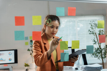 Young businesswoman is writing on a glass wall in the office, using a marker and adhesive notes to brainstorm ideas for a new project