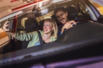 Boyfriend drives the car while girlfriend is taking selfies with him. Happy, young couple.