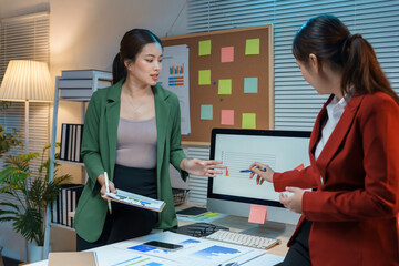 Two businesswomen are discussing over financial data on computer with graphs and charts at night in office