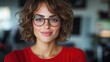 © Dulemegapixel - A young woman adorned in a red sweater and glasses smiles warmly while posing in a modern interior space filled with soft lighting and blurred background.