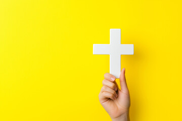 Hand holding a white cross against a bright yellow background, symbolizing faith, hope, and spirituality, perfect for religious, Christian, and inspirational themed designs and concepts.