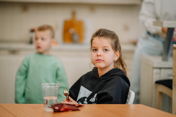 A girl in a black hoodie sits at a table with a small dessert in her hand, while a boy stands in the background..