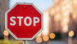 © hardvicore - Stop road sign. Red octagon with white lettering. Blurred city bokeh on background.