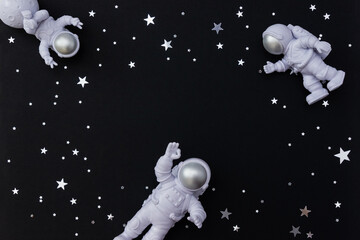 Three astronaut toy figures on black background with silver star confetti. Selective focus, copy space