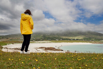 Wall Mural - Teenager girl tourist in yellow jacket standing on a grass field. blue ocean water and white sandy beach in the background. Dog bay, county Galway, Ireland. Stunning Irish nature scene.