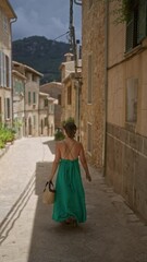 Wall Mural - Woman in green dress walking in valldemossa, mallorca, spain, on a narrow street with traditional stone houses and mountain backdrop under a cloudy sky.