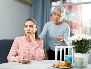 Upset young girl sitting at home table while worried elderly mother reprimanding her. Family conflicts concept