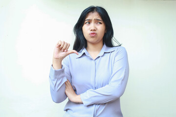 Wall Mural - confused asian young business woman giving thumbs down looking aside with folded arm show upset expression felling dissatisfied wearing formal shirt standing over isolated white background