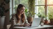 © Ruwan - Remote Work Lifestyle: 'A young woman working from home on a laptop, sitting by a window with plants and cozy decor, showcasing the modern remote work lifestyle with a calm and productive atmosphere.'