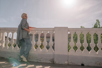 A woman in a gray jacket and blue jeans is leaning against a white railing. She is holding a book in her hand. The scene has a calm and relaxed mood, with the woman enjoying a quiet moment outdoors.