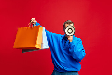 Person, shopper holds two large shopping bags while speaking through megaphone against red background. Sales season. Concept of shopping, Black Friday, marketing, announcement. Ad