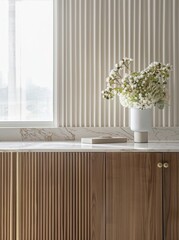 A white vase with white flowers sits on a wooden cabinet with a white marble countertop The cabinet has a fluted design and gold accents A window with white curtains is in the background