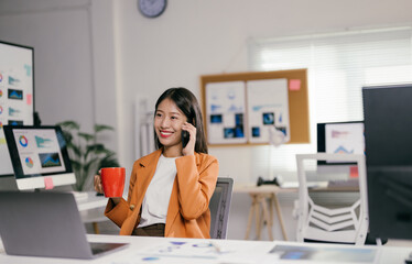 Poster - Asian businesswoman is working in a modern office, drinking coffee and talking on the phone while checking data on her laptop