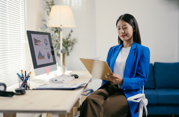 Aufkleber - Young asian businesswoman is sitting at her desk in a modern office, reviewing documents and analyzing data on her computer