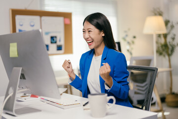 Poster - Young businesswoman is raising her fists in the air, excited about achieving a goal while working at her desk. She is happy with her success