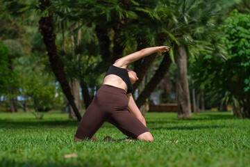 Young woman in sports clothes does yoga in a green park, demonstrating an inclined stretch with her arm outstretched back on a yoga mat surrounded by nature. Concept of smart fitness, yoga, pilates