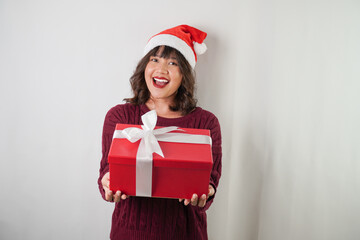 Excited young asian woman wearing santa clause hat and red long sleeved sweater is carrying large gift box with ribbon, isolated over white background. Concept for Christmas Holiday and New Year Party