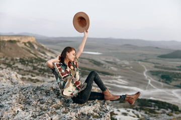 Wall Mural - Serene woman sitting on rock with hat in the air, admiring majestic mountain view