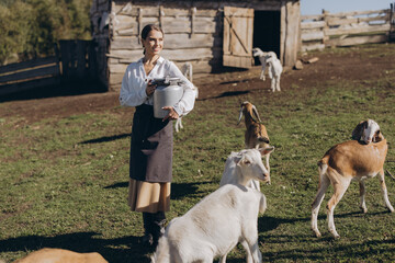 Young Female Farmer Tending to Goats on a Rustic Farm with Wooden Barn on a Sunny Day