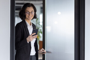 Poster - Businesswoman in office hallway checks smartphone for notifications. Dressed in formal attire, she exudes professionalism while entering through frosted glass door with confident smile.