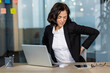 © Liubomir - Businesswoman in office showing signs of back pain while sitting at desk using laptop and phone. Indicates discomfort, poor posture, need for ergonomic solutions in workplace.