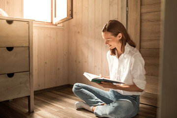 Smiling woman reading book while sitting near window at home, education, knowledge, weekend leisure concept 
