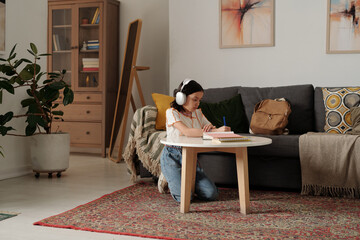 Young girl sitting on floor wearing headphones, sketching in notebook on coffee table in cozy living room with artistic decor and comfortable furnishings