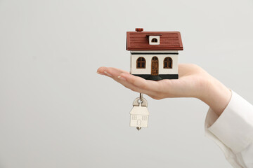 Wall Mural - Woman holding key with keychain and house model on light grey background, closeup. Space for text