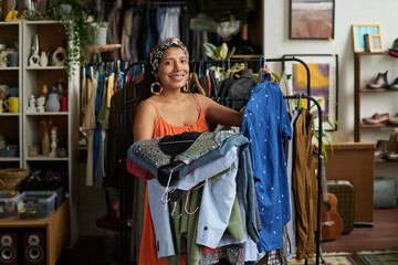 Young cheerful female owner of thrift shop or customer holding stack of casual attire from new collection of secondhand clothes