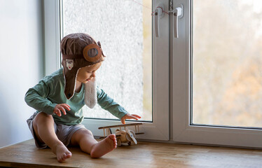 A cute 2-year-old boy in a cap is playing on the windowsill with a wooden airplane. Children's wooden toys for child development.