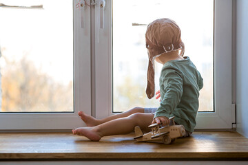 a cute 2-year-old boy in a cap is playing on the windowsill with a wooden airplane. children's woode