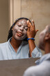 © Roger - A young African woman in a bathrobe applies a face mask while smiling in the mirror.