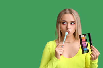 Beautiful young woman with brush and palette of colorful eyeshadows on green background