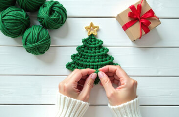 Wall Mural - close-up of hands in a white sweater holding a green knitted Christmas tree with yellow star on a white wooden background, in the background there is a craft gift box with a gift , green yarn